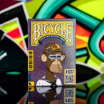 Eine Bicycle Ultimate Bicycle Bored Ape Special Edition Kartenschachtel steht mit sichtbarer Vorderseite vor einem bunten Graffiti-Hintergrund.
