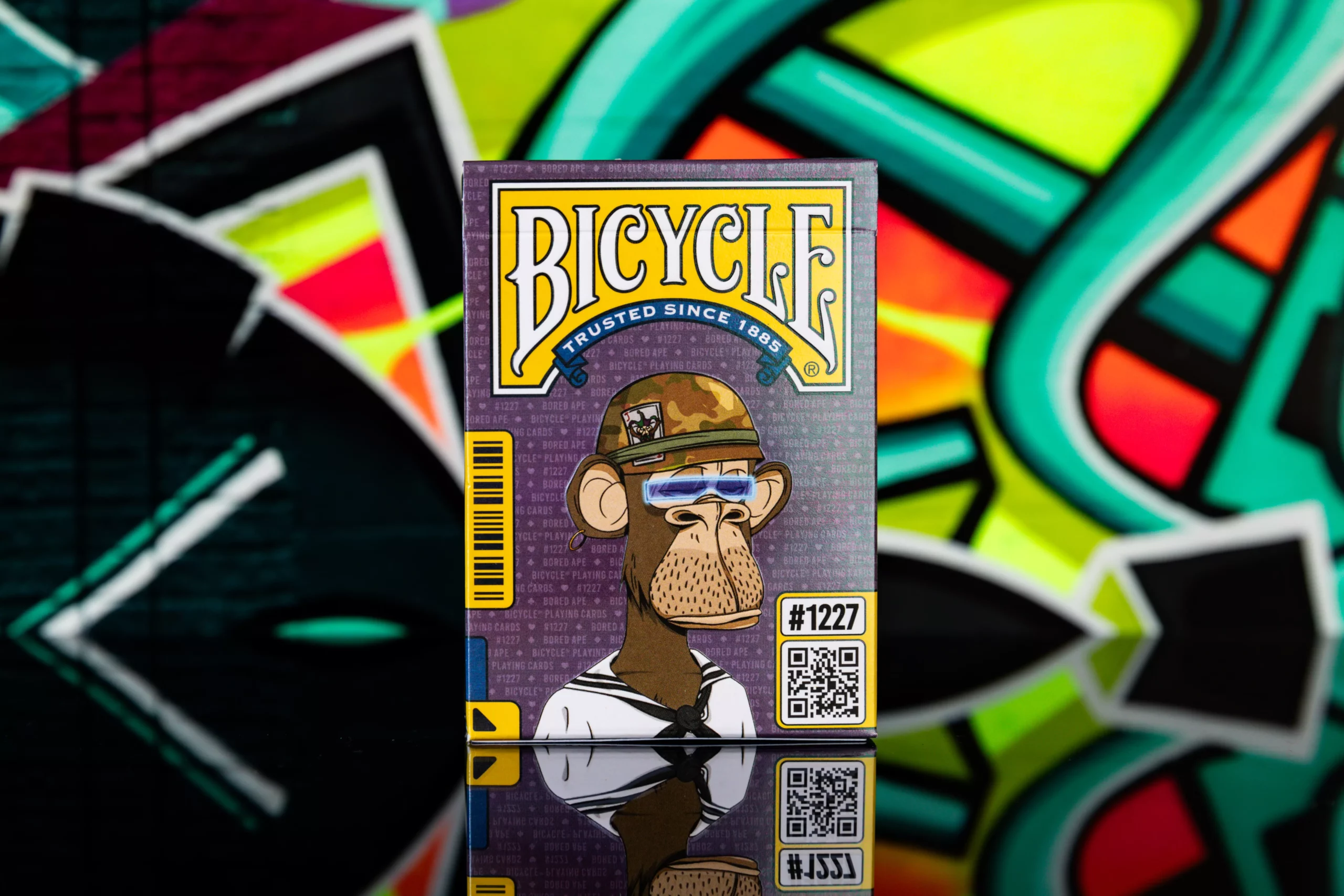Eine Bicycle Ultimate Bicycle Bored Ape Special Edition Kartenschachtel steht mit sichtbarer Vorderseite vor einem bunten Graffiti-Hintergrund.