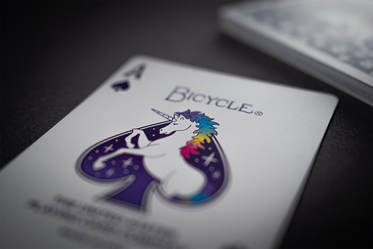 Eine Bicycle Creatives Unicorn Pik Ass Karte liegt vor einem dunklem Hintergrund. Die Illustration der Karte mit Einhorn Motiv ist sichtbar.