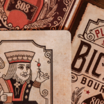 Von einer Bicycle Creatives Bourbon Kartenschachtel ist aus der Nähe 1 Karte zu sehen, im Hintergrund ist eine Rückseite und die Kartenschachtel zu sehen. Die Karte zeigt einen König mit einem Glas Bourbon in der Hand.
