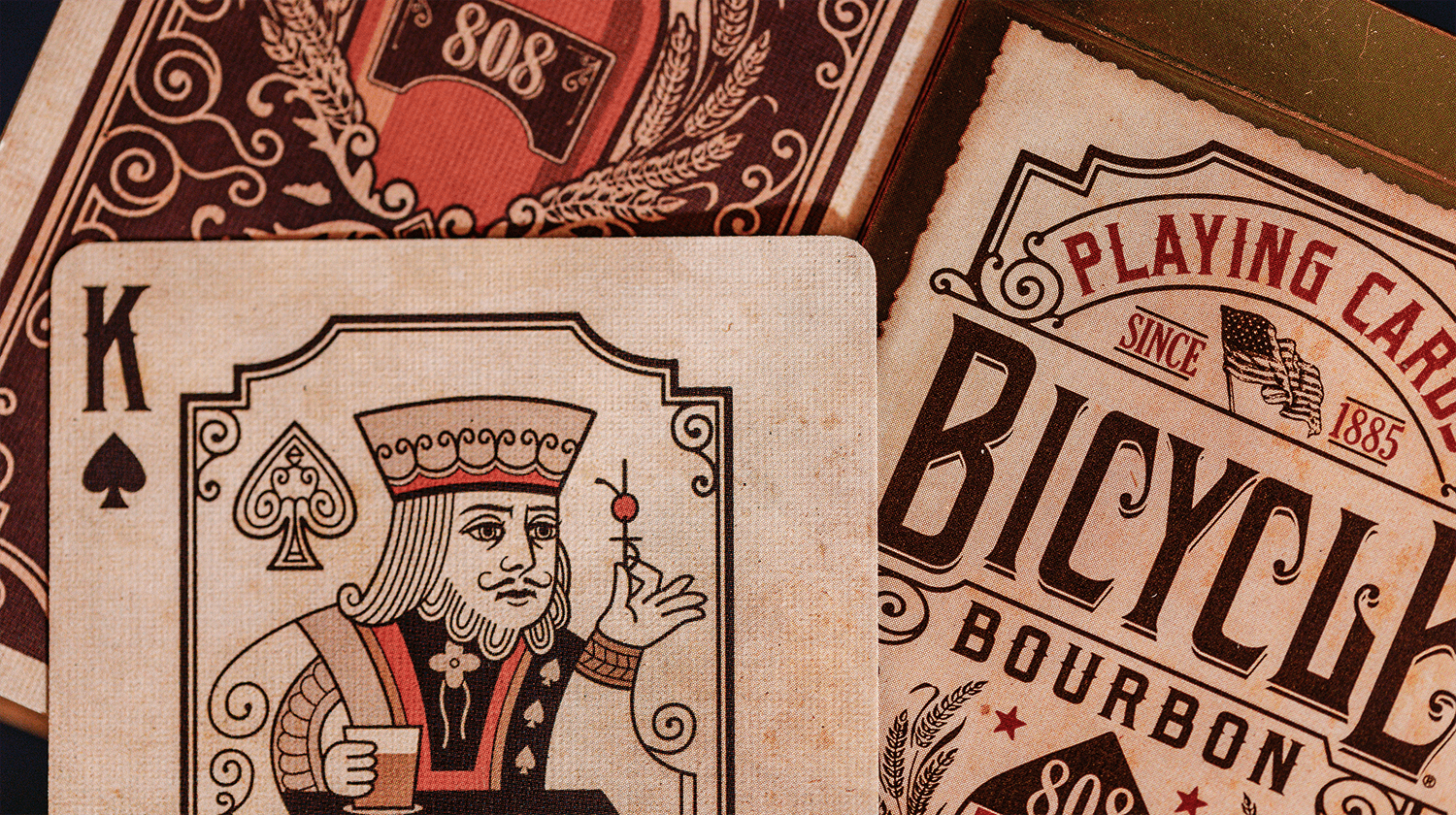 Von einer Bicycle Creatives Bourbon Kartenschachtel ist aus der Nähe 1 Karte zu sehen, im Hintergrund ist eine Rückseite und die Kartenschachtel zu sehen. Die Karte zeigt einen König mit einem Glas Bourbon in der Hand.