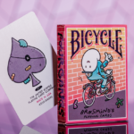 Eine Bicycle Creatives Brosmind Four Gangs Kartenschachtel steht mit sichtbarer Rückseite vor einem violetten Hintergrund aus Backstein, neben der Schachtel schaut eine Karte aus dem Deck hervor.