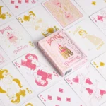 Die Karten einer Bicycle Ultimate Bicycle Disney Princess pink rosa Kartenschachtel liegen ausgebreitet auf einer Fläche sodass man verschieden Vorderseiten und Rückseiten und die Schachtel sehen kann.