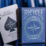 Eine Schachtel aus einem Bicycle Creatives Odyssey Kartendeck steht vor einem blauen Hintergrund, daneben steht ein Ass aus dem Deck.