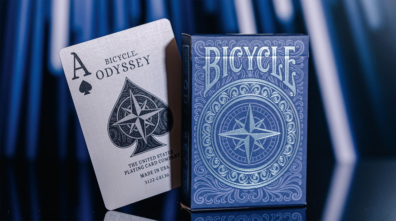 Eine Schachtel aus einem Bicycle Creatives Odyssey Kartendeck steht vor einem blauen Hintergrund, daneben steht ein Ass aus dem Deck.