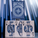 Eine Bicycle Creatives Odyssey Kartenschachtel steht auf einer Erhöhung, darunter stehen drei Karten aus dem deck auf denen man König, Dame und Bube sieht.