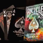 Eine Bicycle Creatives Stargazer Nebula Kartenschachtel steht mit der sichtbaren Vorderseite neben 3 Karten auf denen man die Vorderseite der Karten sehen kann.