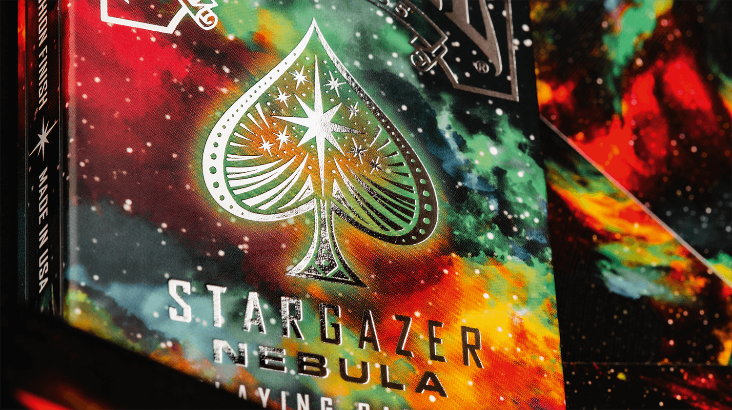Zusehen ist eine Bicycle Creatives Kartenschachtel von Stargazer Nebula in einer nahen Ansicht in der man die Veredelung mit Folie erkennen kann, im Hintergrund sieht man die Rückseiten der Karten.