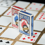 Die Bicycle Trick Deck Blank Back Kartenschachteln stehen auf einem Tisch, darunter ausgebreitet sind Karten aus diesem Deck. Sie haben eine klassische Vorderseite und eine leere Rückseite.