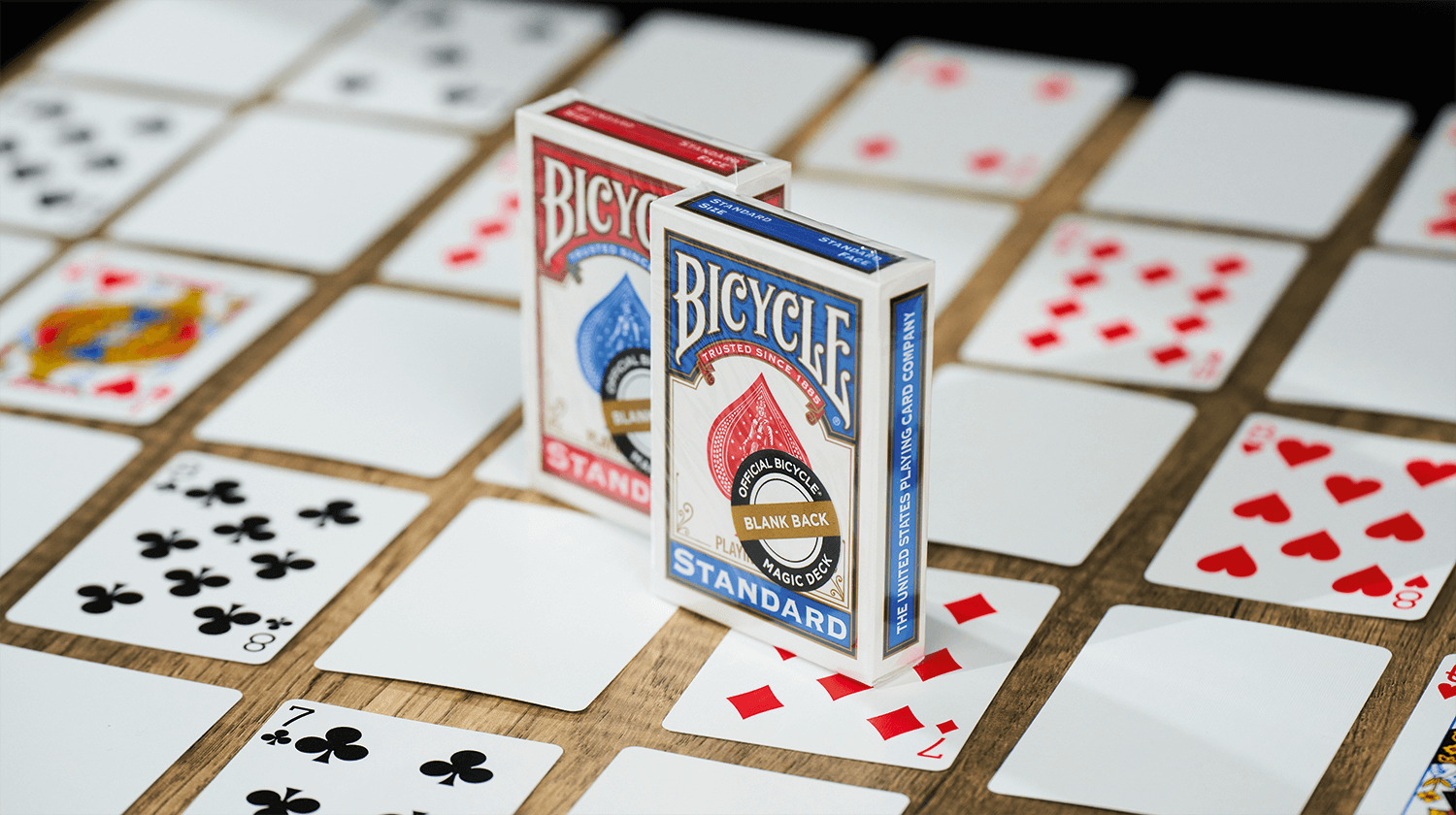 Die Bicycle Trick Deck Blank Back Kartenschachteln stehen auf einem Tisch, darunter ausgebreitet sind Karten aus diesem Deck. Sie haben eine klassische Vorderseite und eine leere Rückseite.