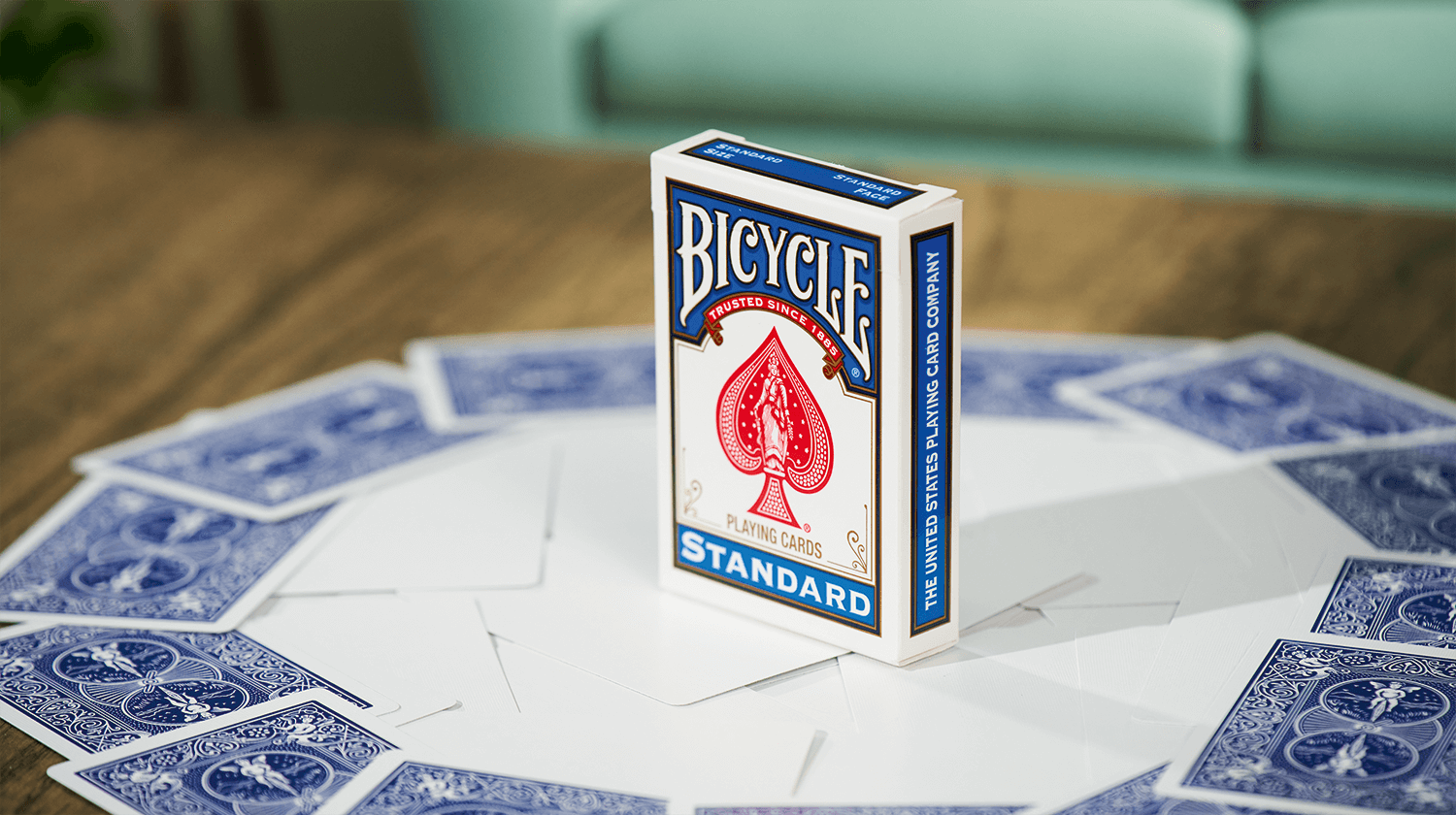 Eine Bicycle Trick Deck Blank Face Blue Blau Kartenschachtel steht auf einem Tisch, unter der Schachtel sind Karten ausgebreitet. Die Karten haben nur eine blaue Rückseite, die Vorderseite ist leer.