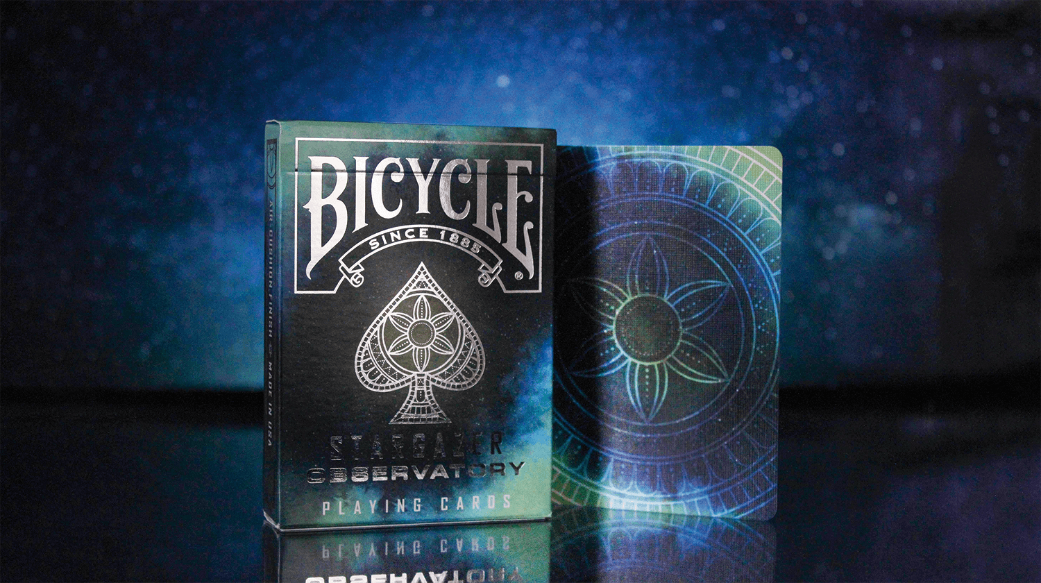 Eine Bicycle Creatives Stargazer Observatory Kartenschachtel steht vor einem Sternenhimmel, hinter der Schachtel ist eine Kartenrückseite zu sehen.