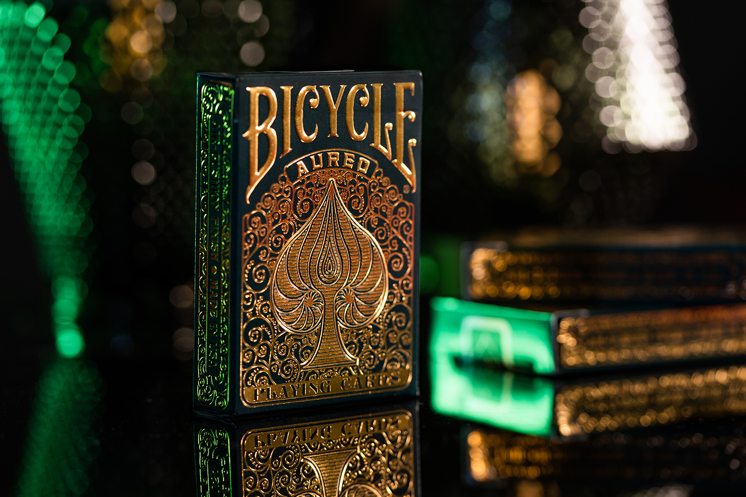Eine Bicycle Creatives Aureo Kartenschachtel steht mit sichtbarer Vorderseite auf einem spiegelndem Untergrund. Der Hintergrund ist unscharf, dunkel und atmosphärisch.