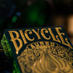 Eine Bicycle Creatives Aureo Kartenschachtel lehnt mit sichtbarer Vorderseite an einer anderen Schachtel vor einem dunklem und unscharfem Hintergrund. Das Bicycle Logo ist sichtbar.