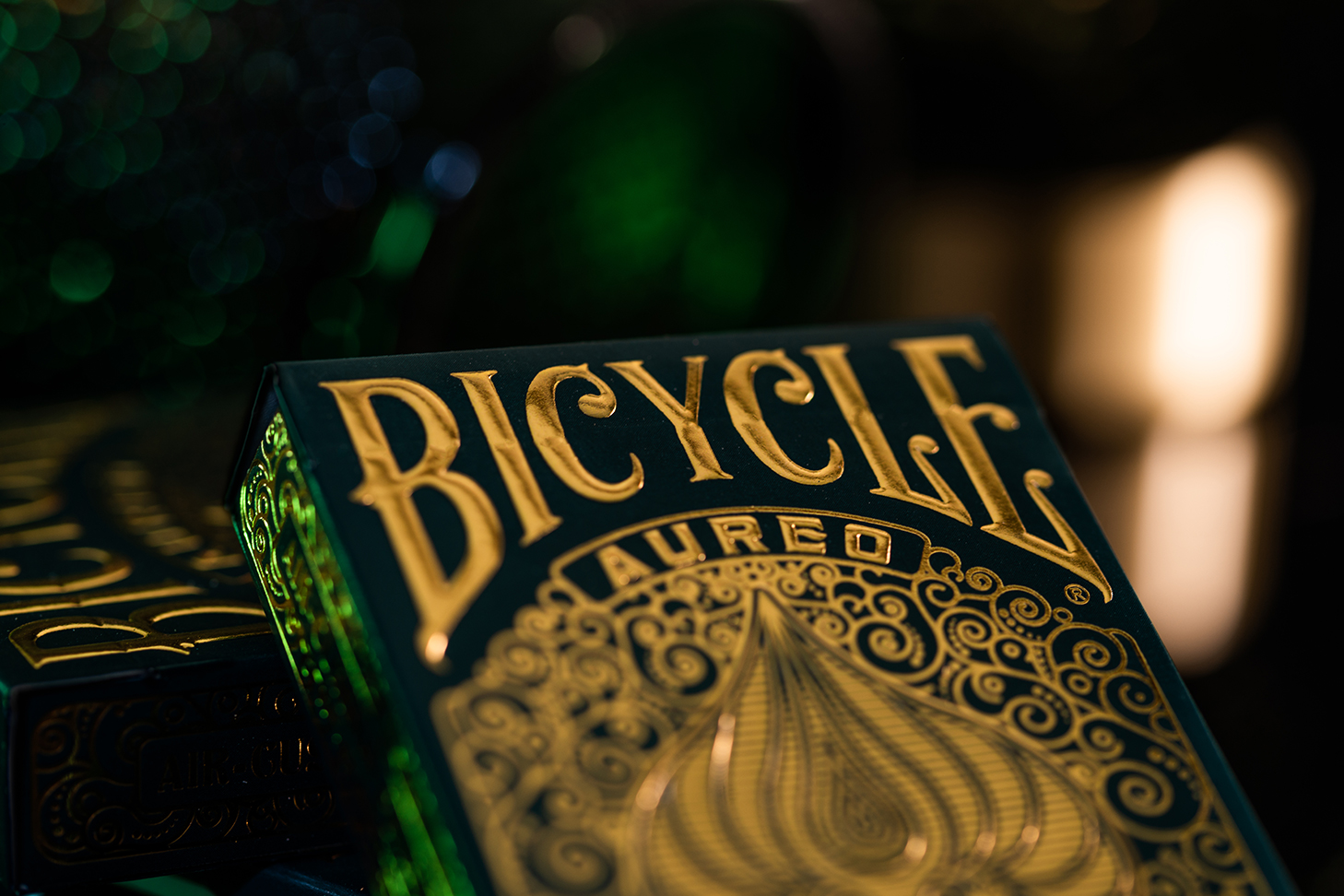 Eine Bicycle Creatives Aureo Kartenschachtel lehnt mit sichtbarer Vorderseite an einer anderen Schachtel vor einem dunklem und unscharfem Hintergrund. Das Bicycle Logo ist sichtbar.