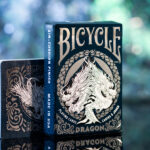 Die Bicycle Creatives Dragon Kartenschachtel mit sichtbarer Vorderseite so wie ein Kartenstapel mit sichtbarem Backdesign stehen vor einem unscharfem Hintergrund.