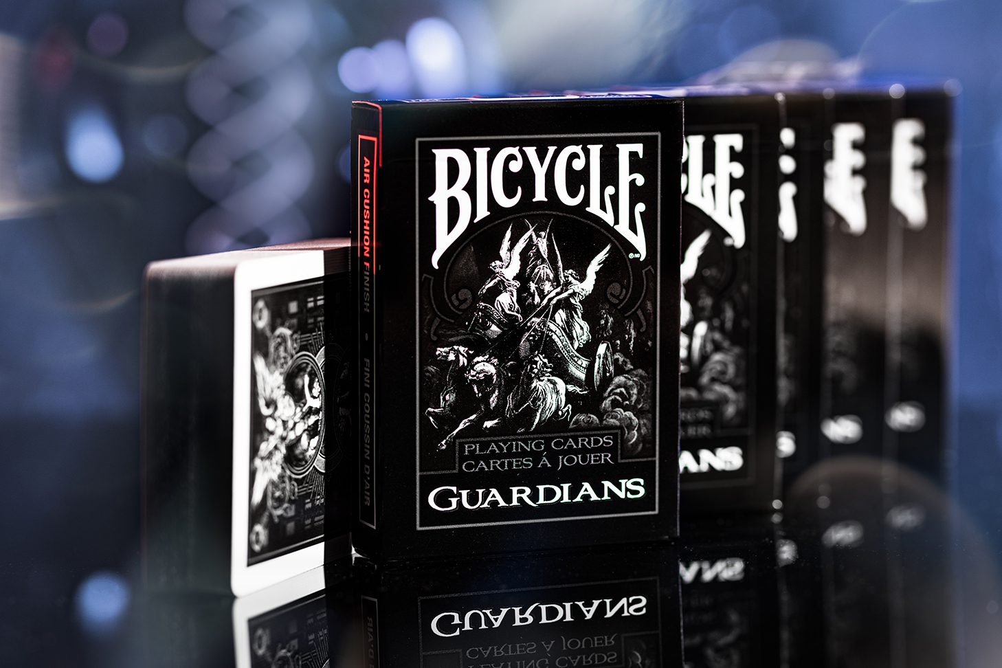 Fünf Bicycle Creatives Guardians Kartenschachteln stehen hintereinander, mit sichtbarer Vorderseite der ersten Schachtel. Daneben liegt ein Kartenstapel mit teilweise sichtbarem Backdesign. Der Hintergrund ist atmosphärisch und unscharf.