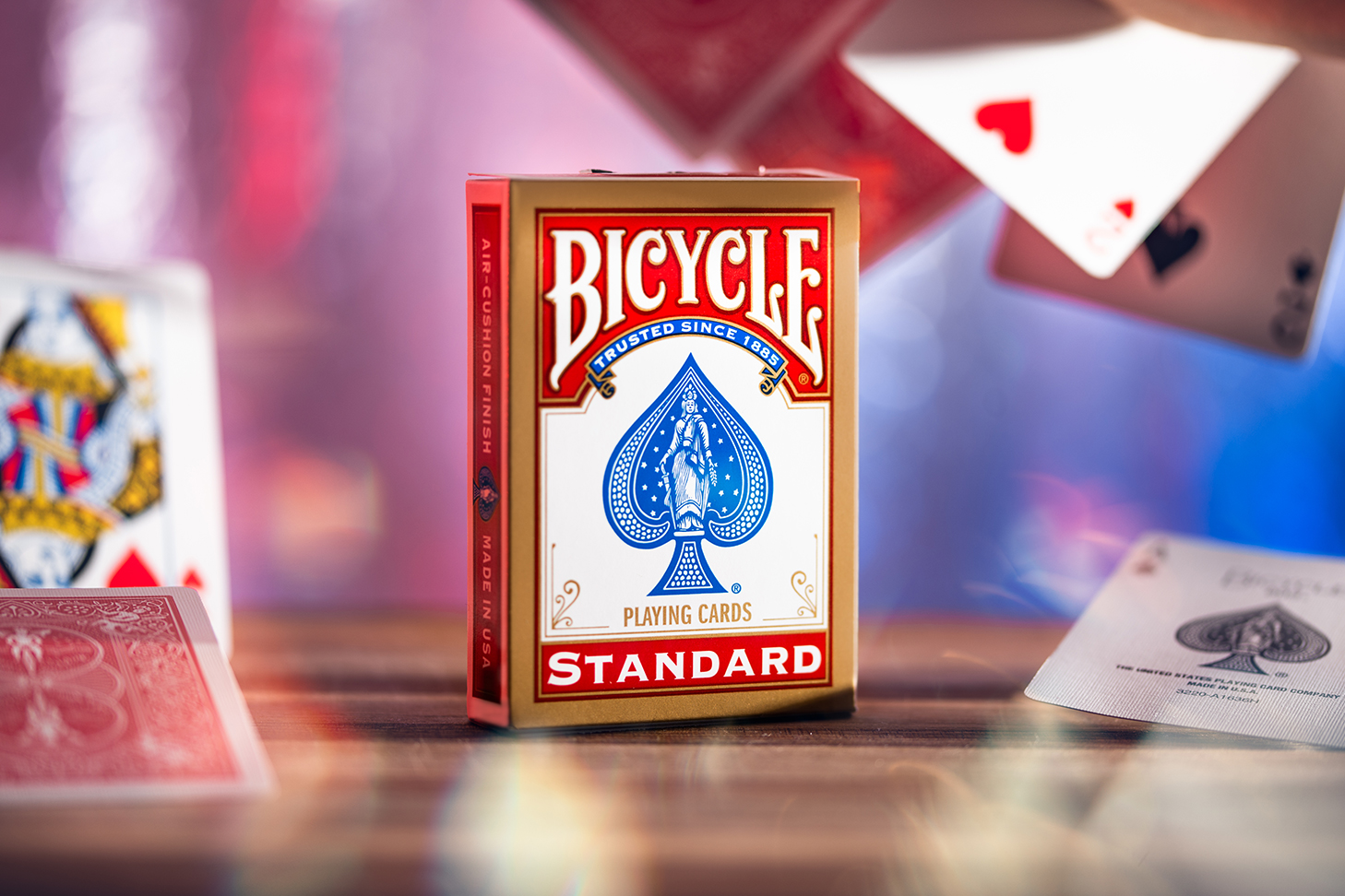 Eine Bicycle Originals Gold Standard Rot Kartenschachtel steht mit sichtbarer Vorderseite vor einem unscharfem Hintergrund. Es fliegen Karten um die Schachtel herum.
