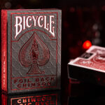Eine Bicycle Ultimates Foil Back Crimson Red Kartenschachtel steht mit sichtbarer Vorderseite vor einem dunklem Hintergrund und wird rot beleuchtet. Eine weitere Schachtel liegt mit sichtbarer Rückseite daneben.