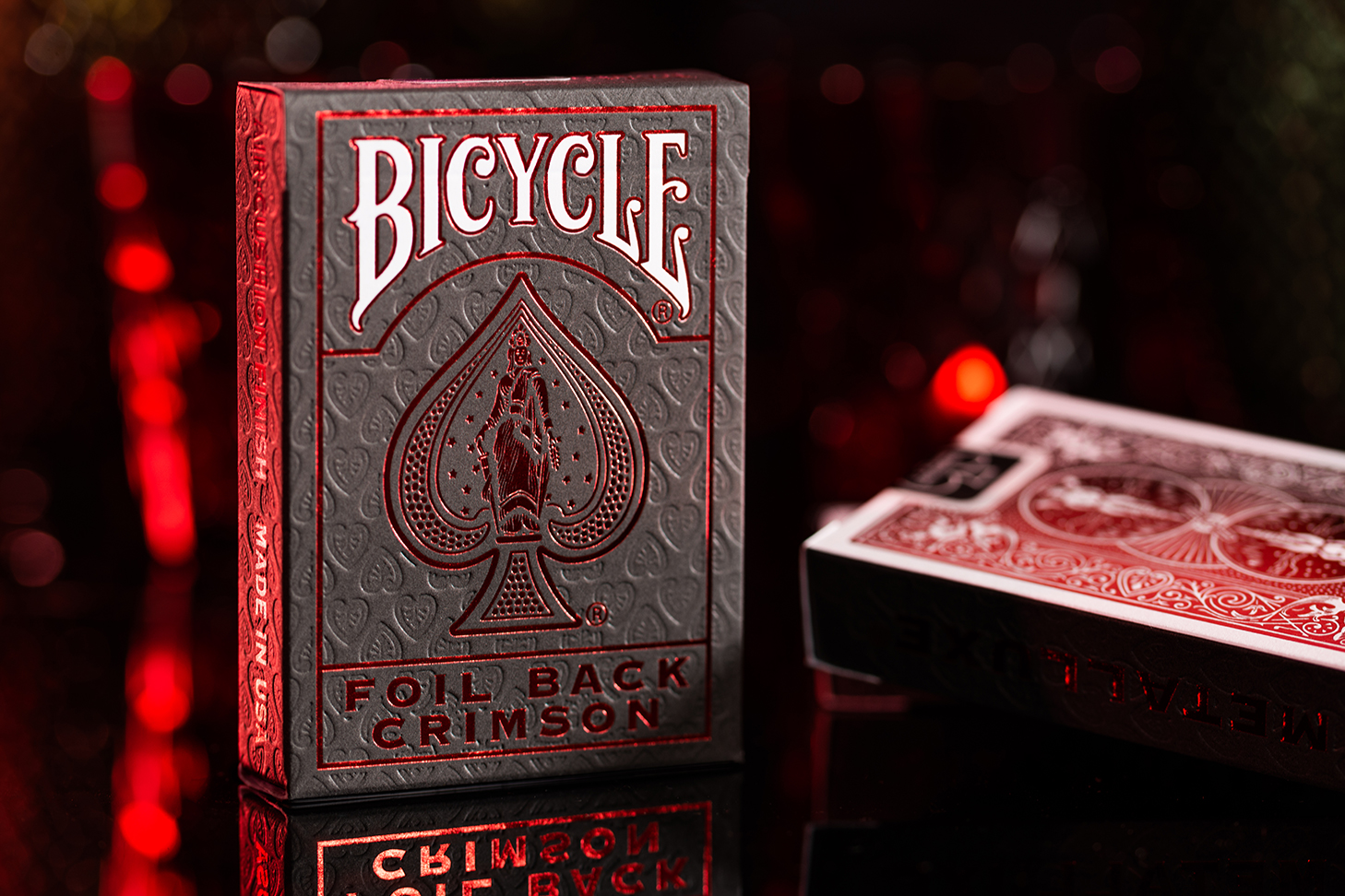 Eine Bicycle Ultimates Foil Back Crimson Red Kartenschachtel steht mit sichtbarer Vorderseite vor einem dunklem Hintergrund und wird rot beleuchtet. Eine weitere Schachtel liegt mit sichtbarer Rückseite daneben.