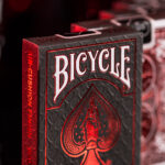 Die Bicycle Ultimates Foil Back Crimson Red Kartenschachtel steht mit sichtbarer Vorderseite vor einem dunklem Hintergrund und wird rot beleuchtet. Die Detailaufnahme ist atmosphärisch und zu einem großen Teil unscharf.