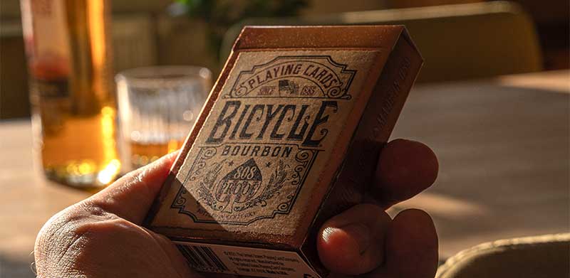Das Kartenspiel Bicycle® Bourbon steht auf einem Tisch vor einem Whisky mit Whisky-Glas. Whisky-Geschenke für Männer, Bicycle Spielkarten