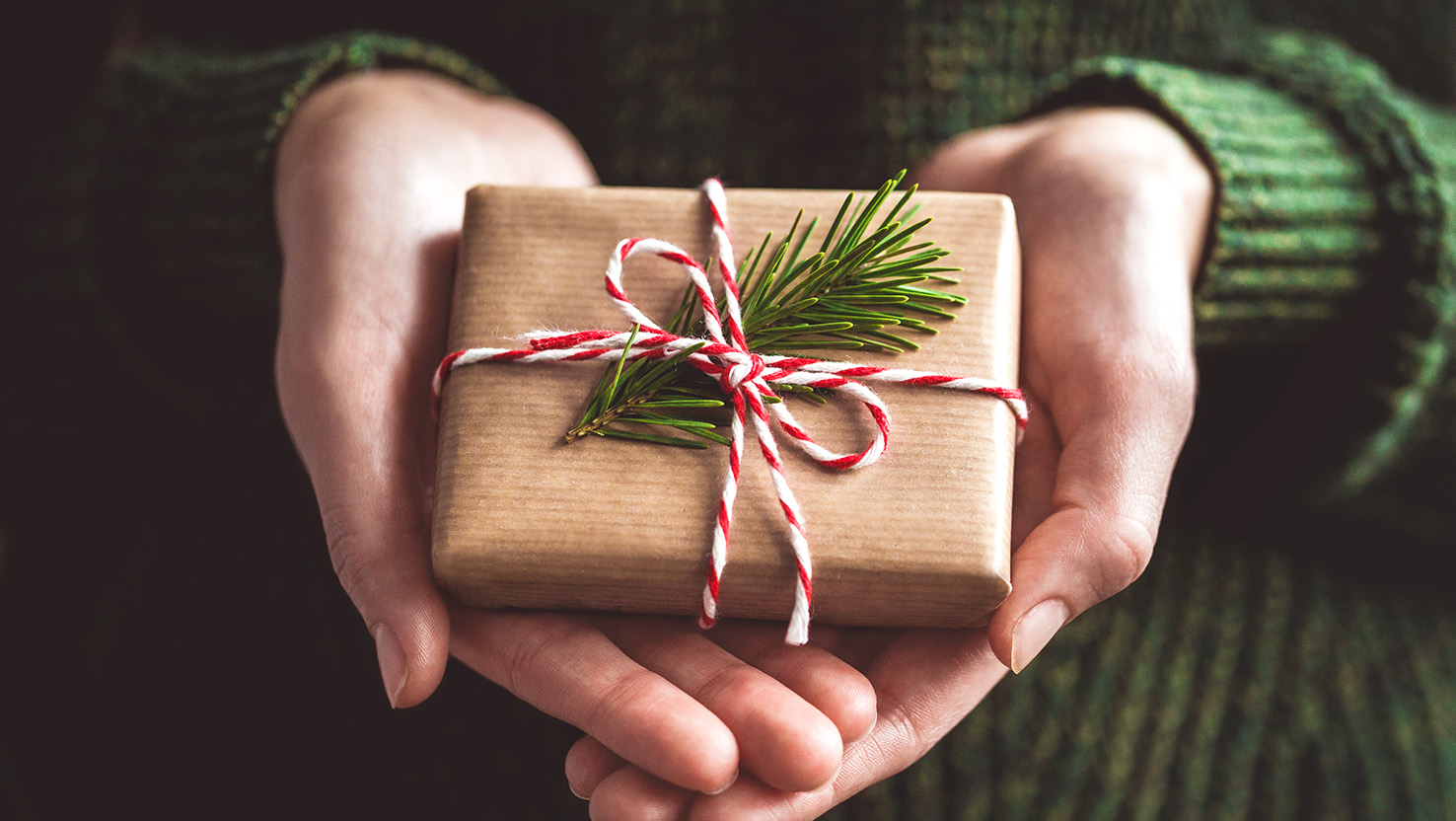 Zwei Hände halten ein Weihnachtsgeschenk, welches in Papier eingepackt und mit einem Tannenzweig und weiß rotem Geschenkband dekoriert ist.