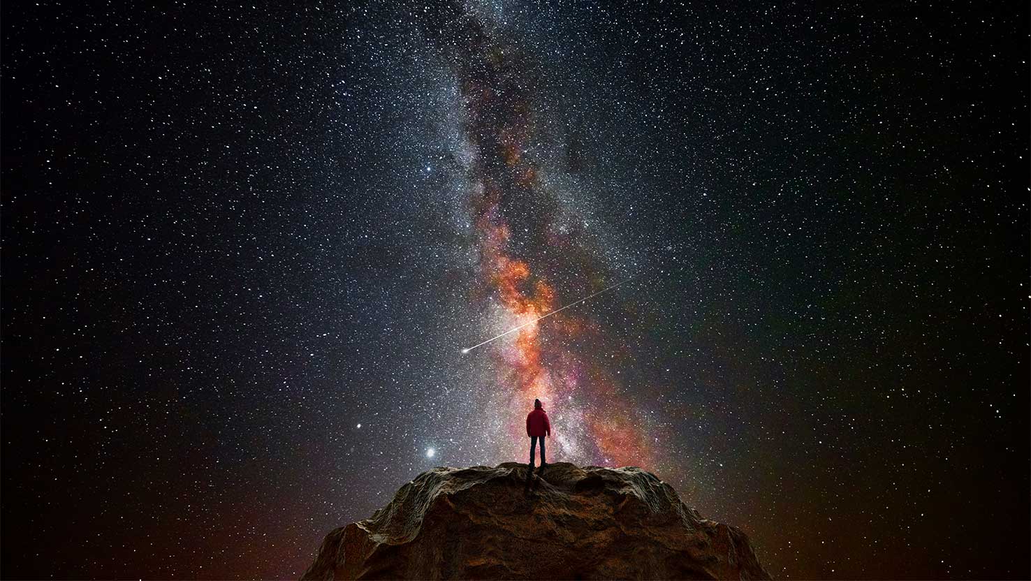 Ein Mensch steht vor dem Sternenhimmel auf einem Fels und sieht die Milchstraße so wie eine Sternschnuppe.