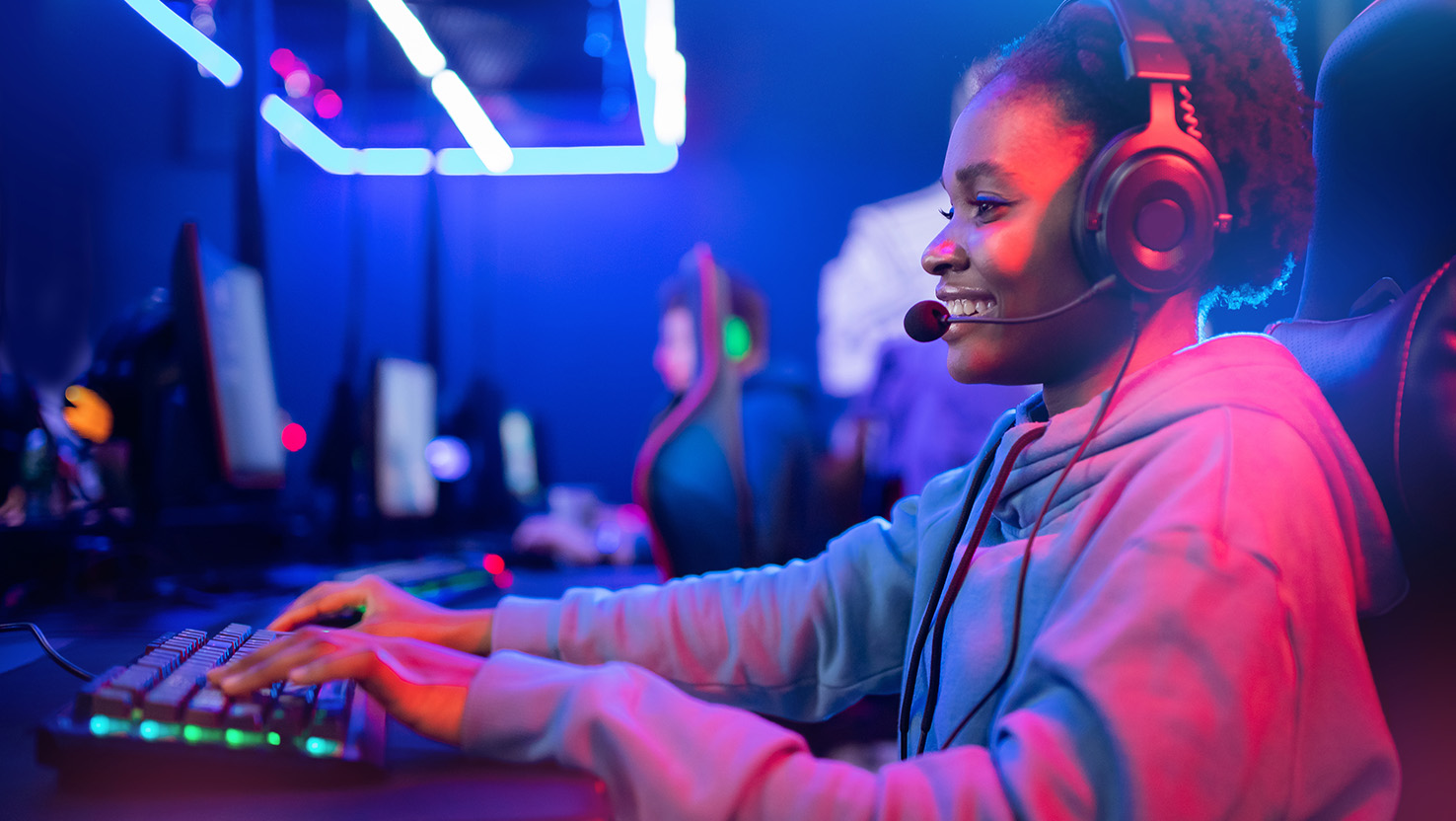 Eine junge Frau sitzt lächelnd mit einem Headset in einem dunklen Raum der von Neonlichtern beleuchtet wird und spielt ein Spiel auf dem Computer.