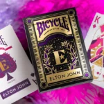 Kartenspiel Bicycle Elton John liegt auf pink-buntem Untergrund neben Pik Ass und Karo König. Elton John Merch, Bicycle Spielkarten